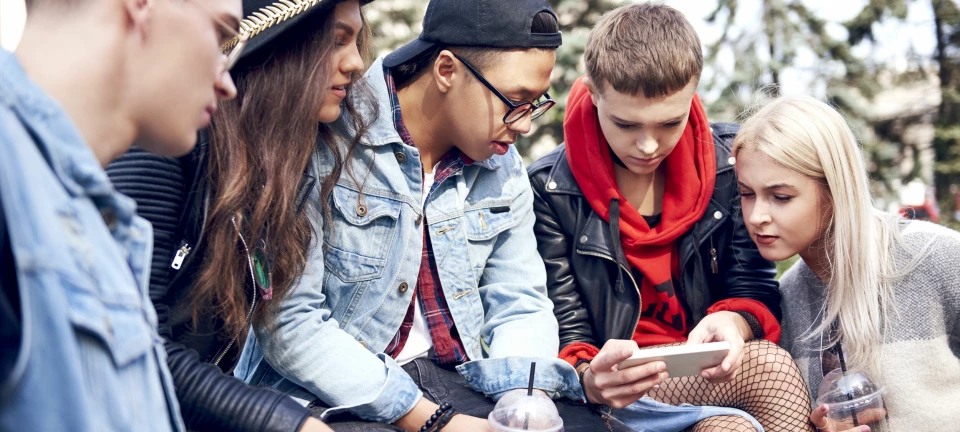 Five young adult friends sitting on wall looking at smartphone in city